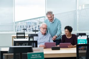 Vrijwilliger van Seniorweb in bibliotheek helpt een man en vrouw die beide een tablet in de handen hebben.